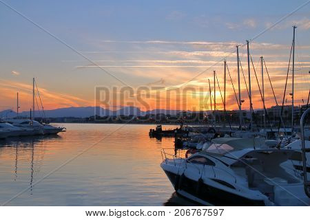The picture was taken in Spain Salou. The picture shows the sun setting over the marina located in the city harbor. You can see the trail of an airplane flying in the sky.