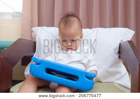 Cute little Asian 18 months / 1 year old baby boy child sitting on sofa watching a video from tablet pc. Kids playing with tablet computer. leisure & children & technology & internet addiction concept