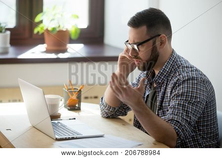 Frustrated male employee discussing contract details over the phone. He is looking at documents in his laptop, holding phone, fling arms in an angry gesture, trying to find solution.