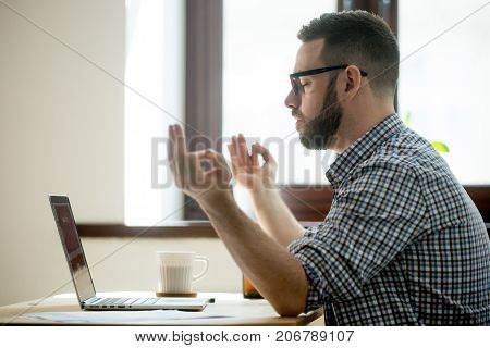 Young adult employee meditating  with hands in zen position, sitting in his office chair. Breathing exercise to build up concentration, cope with stress. Stress free life, mindful lifestyle concept