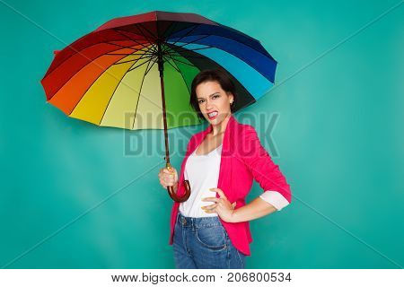 Agressive stylish girl in bright casual clothes under rainbow colored umbrella posing at azur studio background, copy space