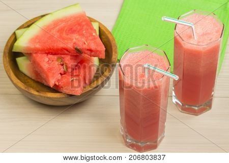 Watermelon juice in two glass tumblers with straw on a light wooden background, a delicious cocktail, a plate with slices of ripe watermelon on the table with a green cloth