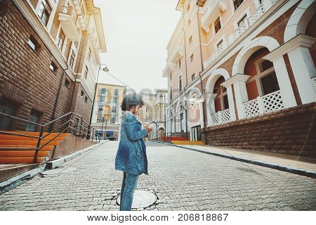 Biracial young woman with curly hair is standing on pavement stones of empty urban street and operating white flying drone using remote device with screen joystick and antennas; sunny autumn day