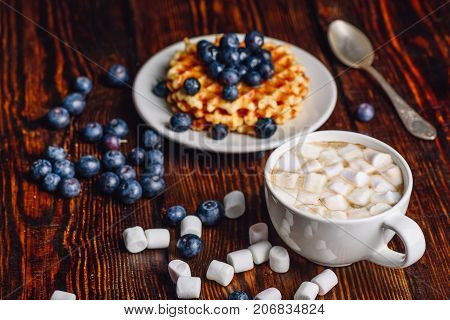 Cup of Hot Cocoa with Marshmallow with Belgian Waffles on Plate with Fresh Blueberry. Spoon and Some Berries and Marshmallows Scattered on Wooden Table.