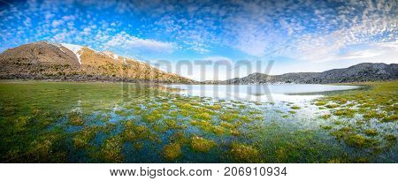 Amazing panorama of Omalos plateau with lake, snowy mountains, grass, cloudy blue sky, Lasithi, Crete