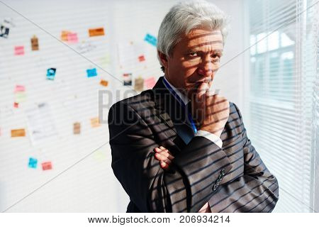 Pensive mature FBI agent rubbing his chin while thinking over committed crime, investigation board with collected evidence on background, portrait shot