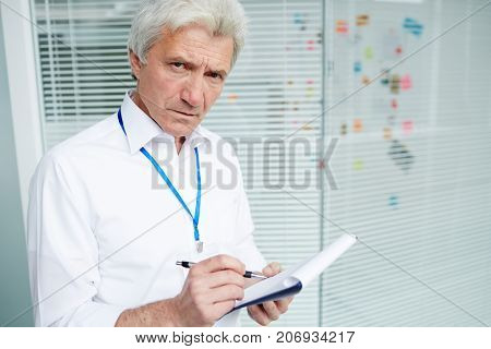 Waist-up portrait of confident senior police officer looking at camera while standing at open plan office and analyzing results of expert study, he holding clipboard in hands