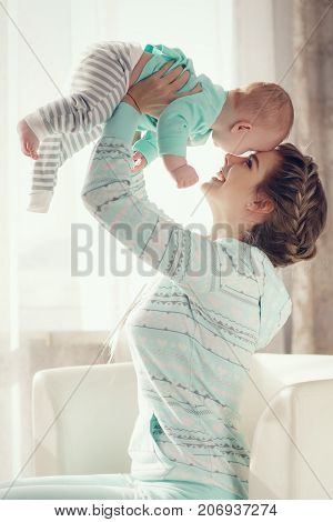 Happy young woman with thick blond hair braided in a thick braid,a pleasant smile,dressed in a light green track suit,playing with his newborn son in the bedroom on the bed,boy with short,fluffy hair,light green shirt and striped pants