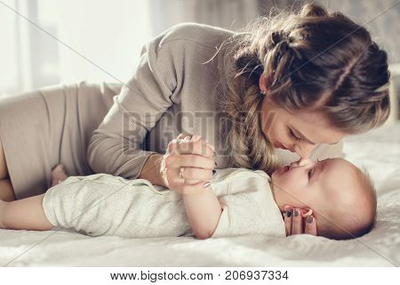 Beautiful young woman with thick blond hair braided in a thick braid,makeup and a nice smile, wearing a wedding ring and earrings,wearing a dress of beige color,playing with his newborn son in the bedroom on the bed,boy with short,fluffy hair