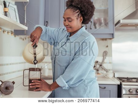Smiling young African woman standing alone at her kitchen counter preparing fresh coffee by pouring hot water into a french press