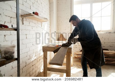 Skilled carpenter with handsaw cutting wooden plank in workshop interior. Young craftsman holding arm saw sawing board on workbench table, using woodworking tools. Carpentry and construction concept