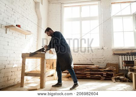 Young trained carpenter working in his carpentry shop, woodworker cutting wooden plank with handsaw for custom-made furniture. Skilled craftsman sawing wood board on table using portable arm saw