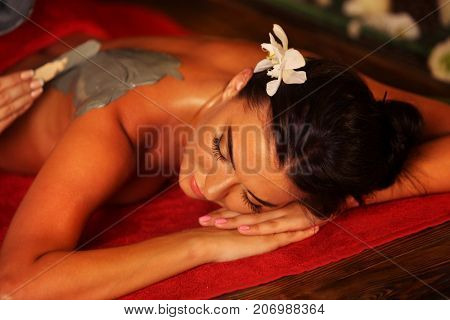 Mud mask of woman in spa salon. Girl on Luxary interior with candles in oriental therapy room. Top view of female with flower lying on wooden spa bed. Rest in an elite beauty salon.