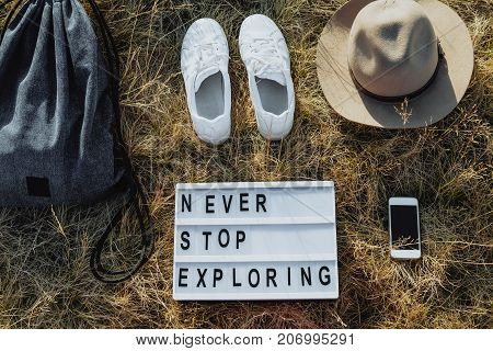 Flat lay travel composition with hat backpack white sneakers phone board with text Never stop exploring on dry grass Top view