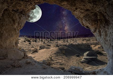 An unrealistic night landscape through a cave with milky, moon in eclipse, and desert with ruins of ancient columns.