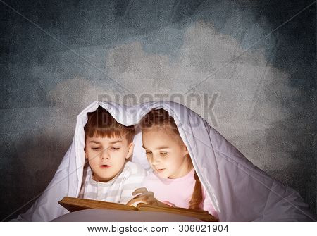 Engrossed Little Girl And Boy Reading Book In Bed After Bedtime. Pretty Kids Together Hiding Under B