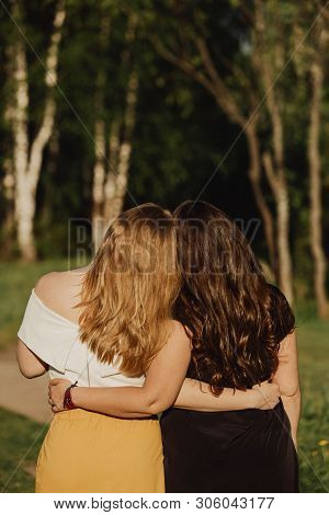 Young Women Different Build With Long Hair Are Embracing On The Green Field In The Evening. Friendsh
