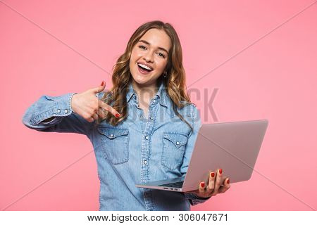 Happy blonde woman wearing in denim shirt holding laptop computer and pointing on it while looking at the camera over pink background