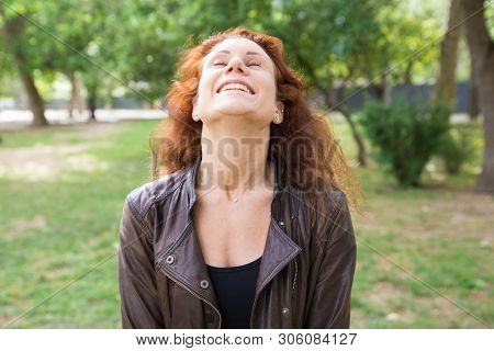 Happy Pretty Young Lady Throwing Head Back In City Park. Woman Wearing Leather Jacket And Keeping Ey