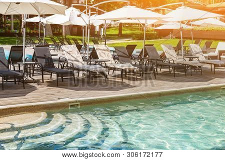 Poolside With Clear Blue Water And Loungers Under Umbrellas On Summer Day Heat. Swimming Pool At Res