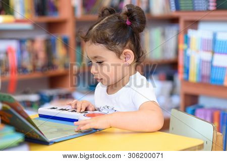 Little Child Indoors In Front Of Books. Cute Young Toddler Sitting On A Chair Near Table And Play Wi