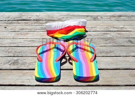 closeup of a pair of rainbow-patterned flip-flops and a rainbow-patterned swimsuit on a weathered dock, next to the ocean or a pond