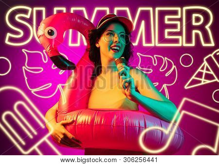 Female Model In Neon Light On Purple Background. Modern Design. Beautiful Young Woman In Cap And Sun