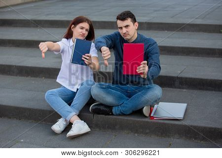 University Students With Thumb Down Gesture Outdoors On Campus Sitting On Stairs With Books In Hand.