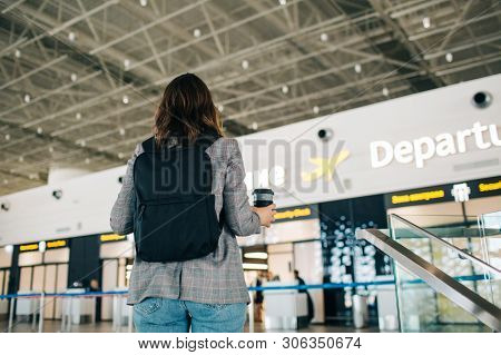 Young Girl Stands In Front Of Departure Area At The Airpot With Her Backpack And Coffee Cup.  View F