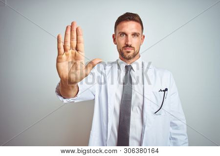 Young handsome doctor man wearing white profressional coat over isolated background doing stop sing with palm of the hand. Warning expression with negative and serious gesture on the face.