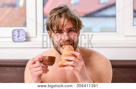 Guy Having Coffee And Holds Croissant While Lay Bed In Bedroom Or Hotel Room. Bearded Man In Bed Dri
