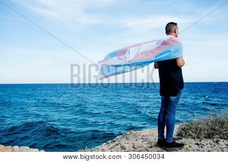 a young caucasian person, seen from behind, draping a transgender pride flag over his or her shoulders, facing the ocean