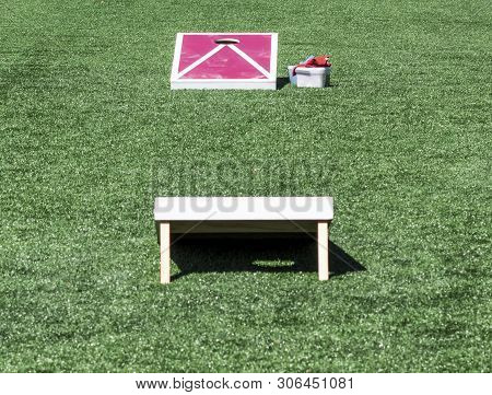 A Green Turf Field Has Two Red And White Corn Hole Games Set Up Ready To Play.