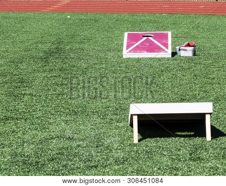 Two Red And White Corn Hole Boards Are Set Up On A Green Turf Field Across From Each Other.