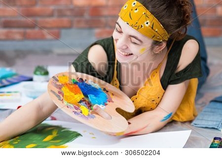 Home Art Studio. Portrait Of Happy Female Painter Lying Down On Floor, Holding Palette, Creating Art