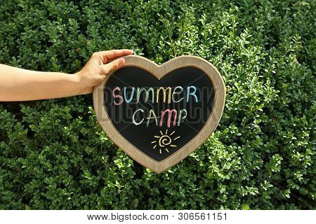 Woman Holding Heart Shaped Blackboard With Text Summer Camp And Drawing Near Green Bush, Outdoors