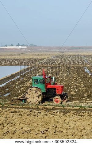North Korea - May 4, 2019: Countryside Scene. Tractor With A Red Flag On The Cab Plowing Agricultura