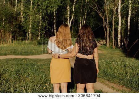 Young Women Different Build With Long Hair Are Embracing On The Green Field In The Evening. Friendsh