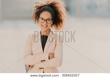 Confident Cheerful Female Entrepreneur With Curly Afro Hair, Keeps Arms Folded, Uses Modern Tablet C