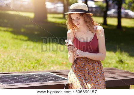 Horizontal Short Of Young Girl Charges Mobile Phone Via Usb Outdoors, Lady Sits On Bench With Solar 