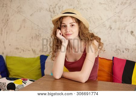 Indoor Image Of Attractive Positive Female Sitting On Sofa, Putting Her Arms On Table, Touching Face
