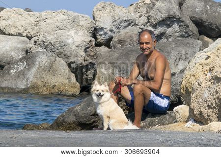 Cyprus, Karavas, Alsancak - June 10, 2019: Adult Male Cypriot Sitting With A Cute Spitz Dog On Backg