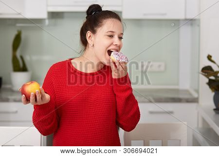 Pregnant Woman In Kitchen Holding Apple In Hand And Eating Cake. Young Attractive Female Decides To 