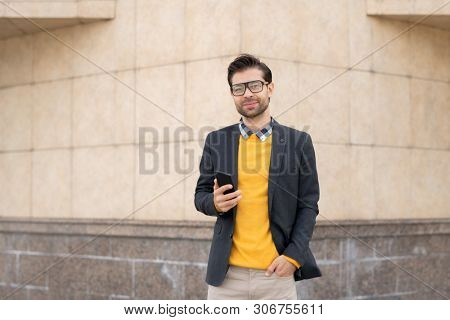 Happy young smart businessman with smartphone messaging while standing by wall of modern architecture in front of camera