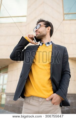 Young elegant man in yellow pullover and dark blue jacket talking on mobile phone against modern architecture