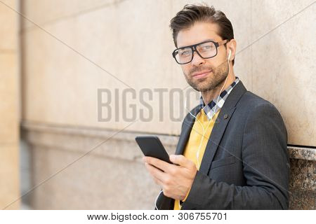 Young man in smart casualwear, eyeglasses and earphones listening to music from playlist in smartphone outdoors
