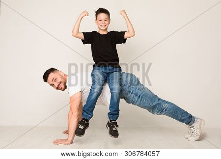 Family Of Strongmen. Dad Doing Push Ups With Son On Back, Cheerful Boy Showing Biceps On Camera