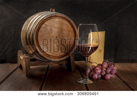 A Glass Of Red Wine With A Wine Barrel, A Large Piece Of Cheesse, And Grapes On A Dark Rustic Backgr