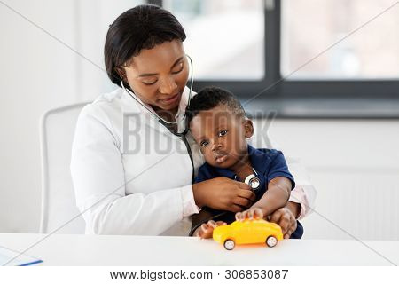 medicine, healtcare, pediatry and people concept - african american female doctor or pediatrician with stethoscope listening to baby patient on medical exam at clinic