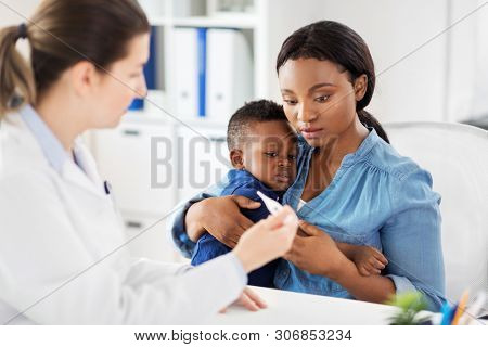 medicine, healthcare and pediatry concept - african american mother with sick baby son and caucasian doctor with thermometer at clinic
